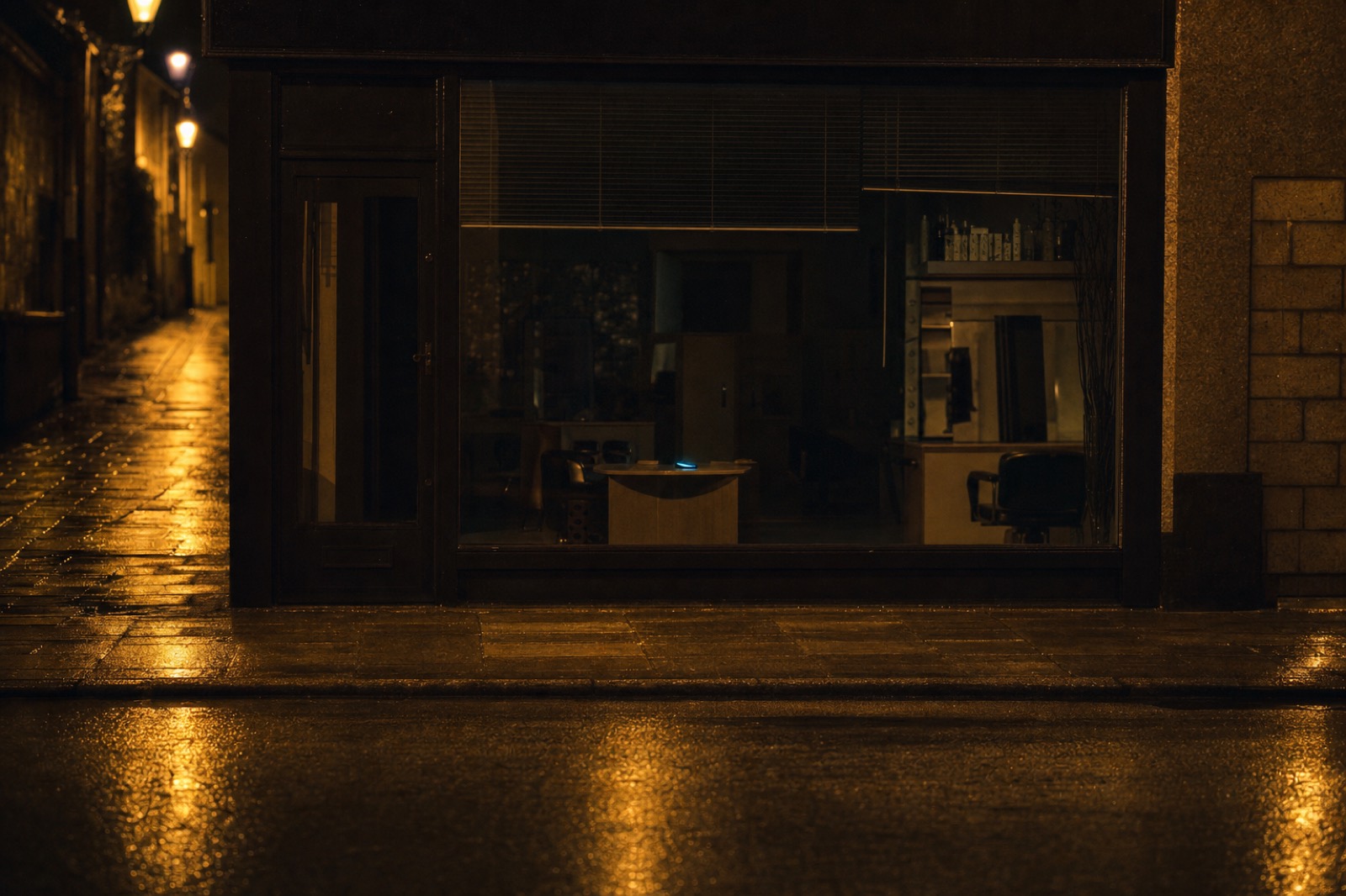 A closed salon at night on a wet British high street, one phone glowing on the counter inside.